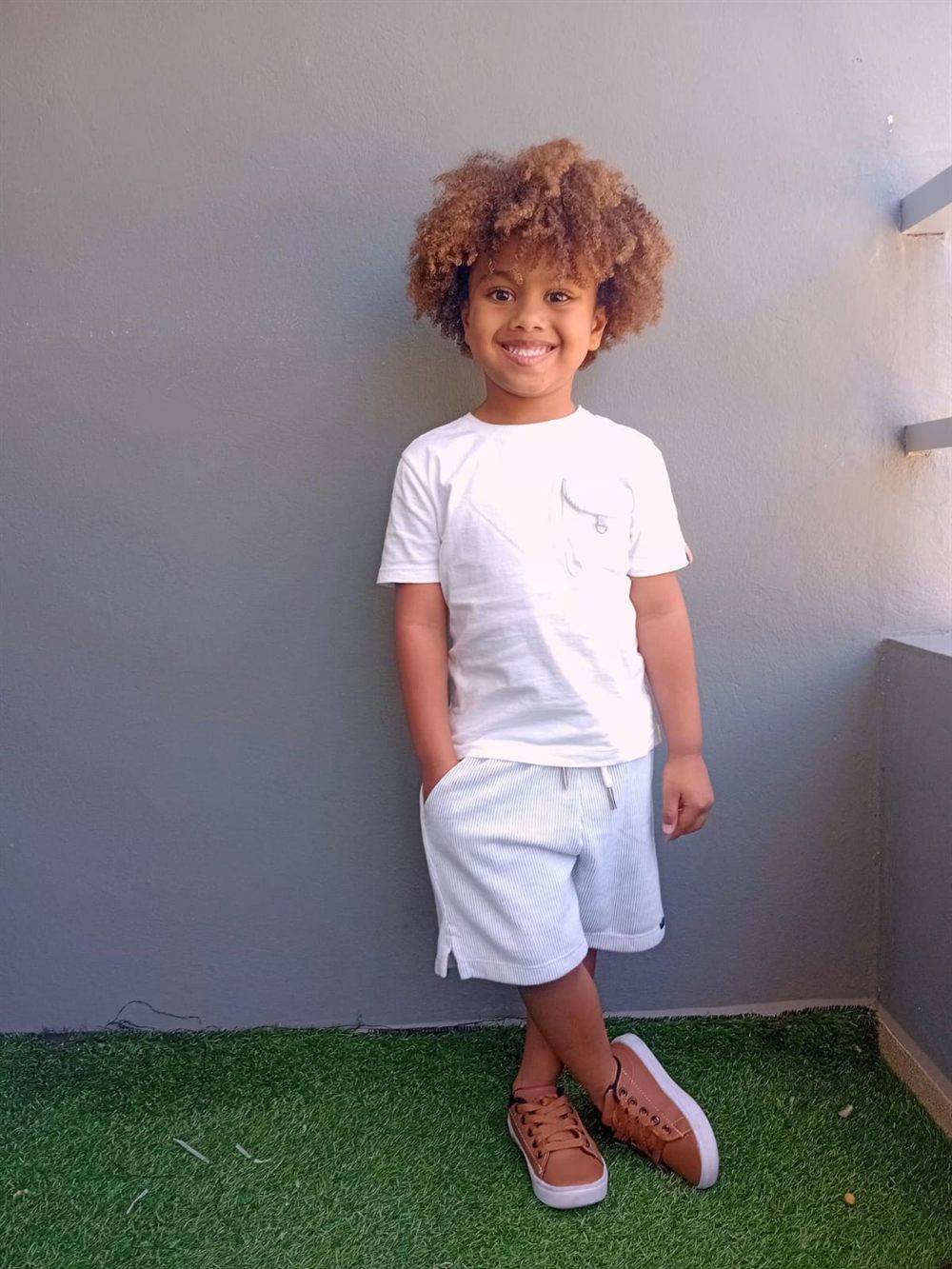 Isaac Roberto de camiseta branca e bermuda listrada clara, encostado numa parede cinza