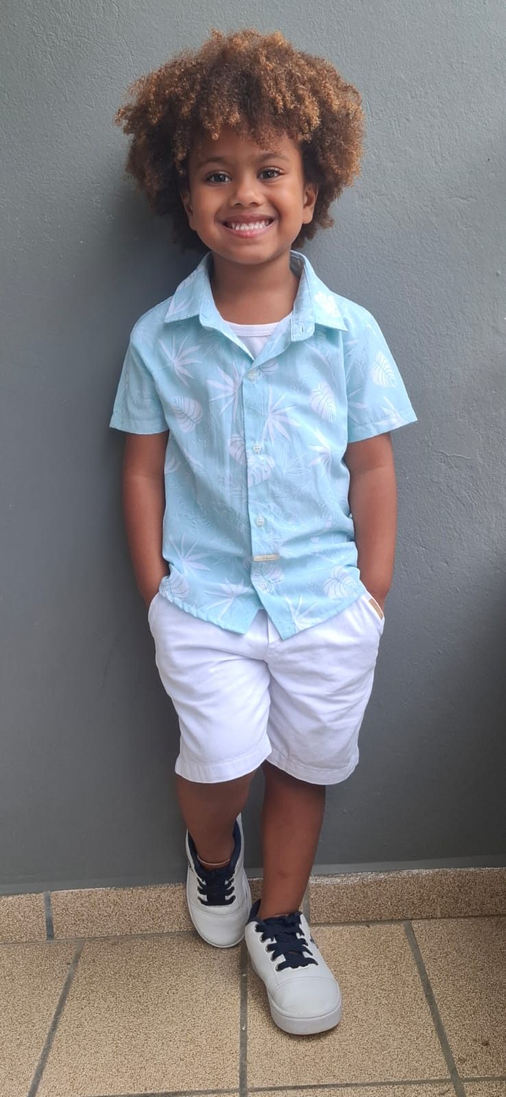 Isaac Roberto de camisa floral azul-clara e bermuda branca, encostado numa parede cinza